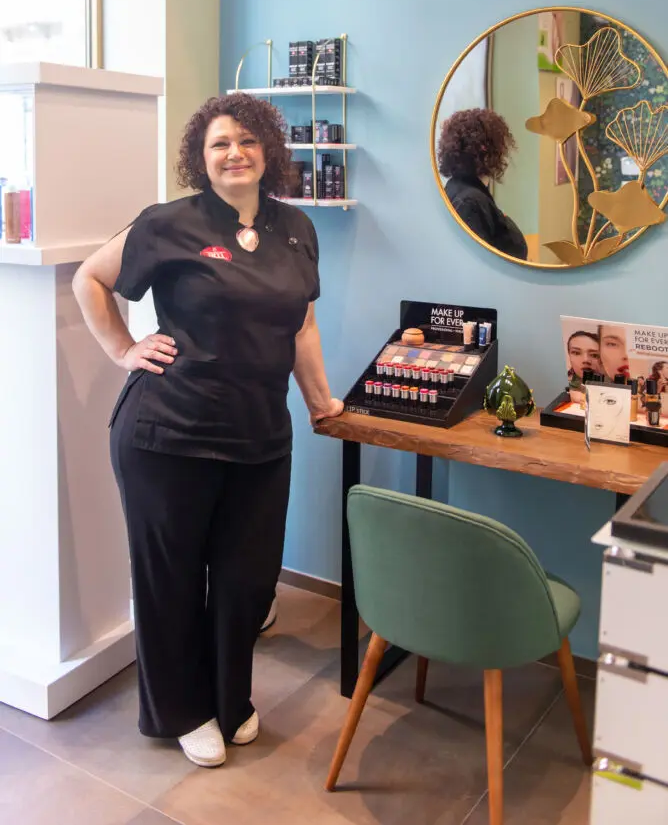 Alessandra Marulli in divisa nera accanto a un banco in legno con espositore Make Up For Ever e sedia di velluto verde