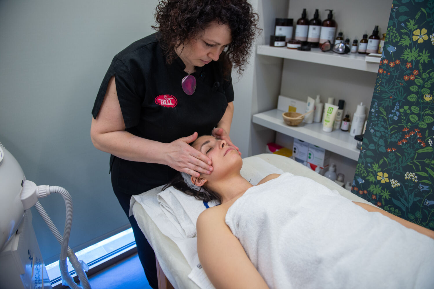 Estetista A.M. Estetica con divisa nera massaggia il viso di una cliente avvolta in un telo bianco sul lettino
